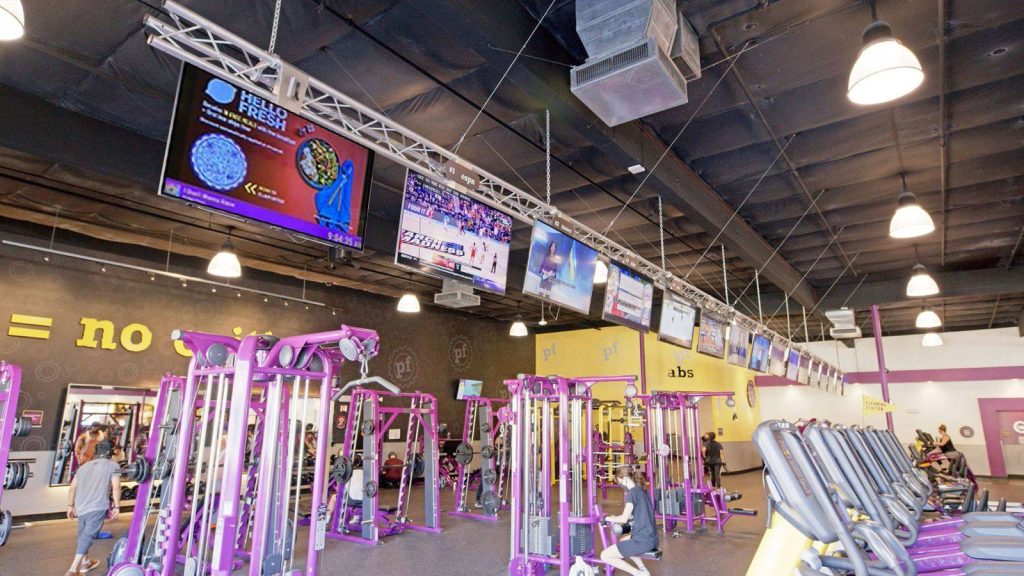 Row of ceiling-mounted screens above a Planet Fitness gym floor, one running a HelloFresh ad, with members using equipment below