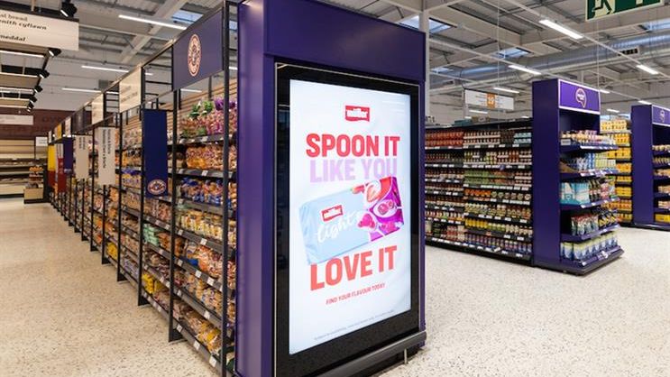 Large purple digital screen at the end of a supermarket aisle running a Müller Light campaign, surrounded by stocked shelves