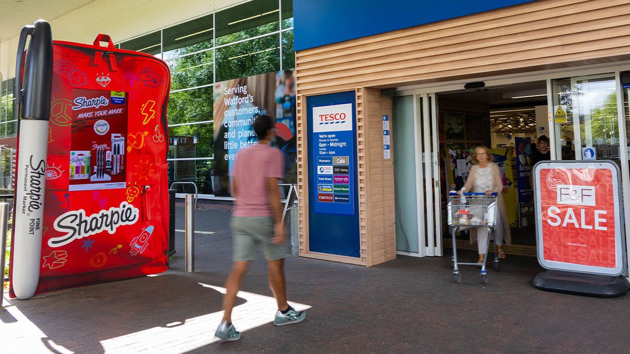 Shoppers entering a Tesco supermarket past a giant inflatable Sharpie display and an F&F sale sign at the store entrance